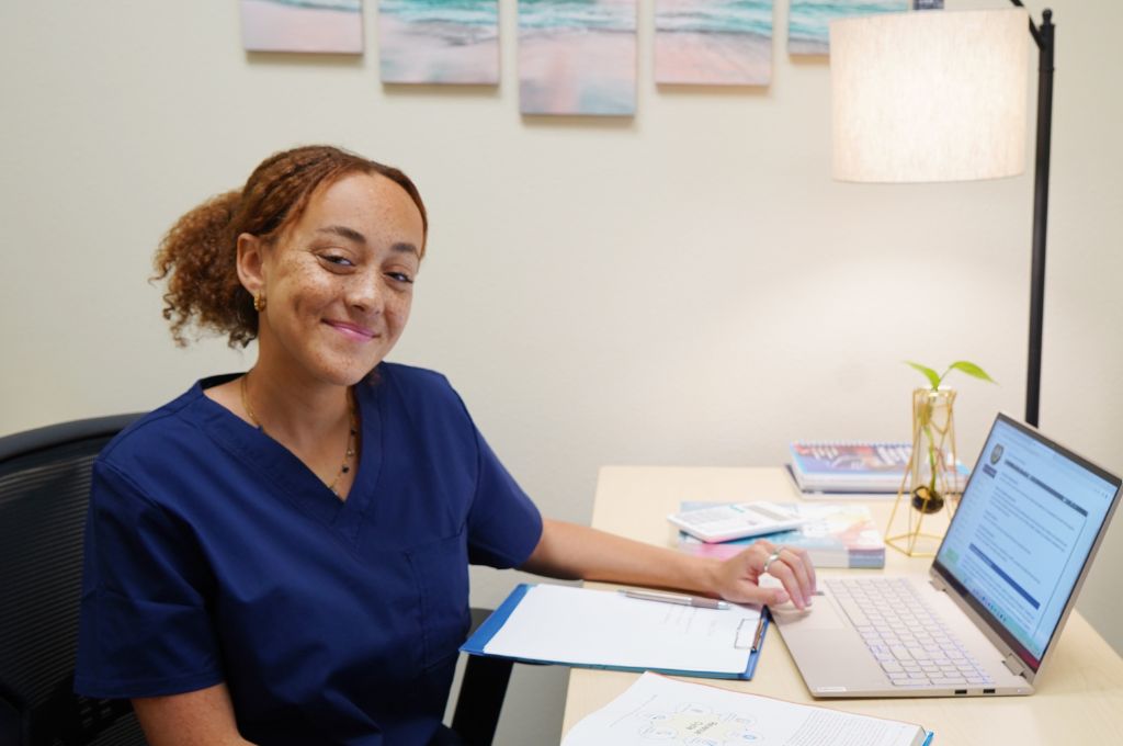 A medical billing and coding specialist in blue scrubs working at a desk with a laptop and paperwork. The professional is reviewing documents with a warm smile, representing a career in healthcare administration.