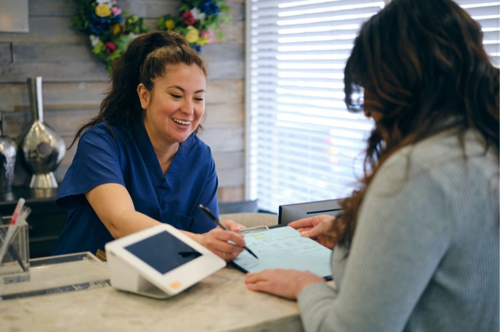 A medical billing and coding professional in blue scrubs assisting a patient with paperwork at a front desk. The healthcare worker is smiling while helping the patient complete necessary forms.