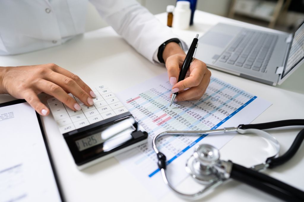 A medical billing and coding specialist analyzing healthcare data with a pen and calculator. A laptop, medical charts, and a stethoscope are on the desk, representing the intersection of healthcare and administrative work.