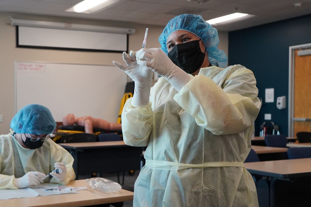 Pharmacy technician students in Texas practicing sterile compounding techniques as part of their certification training. A student in protective gear, including a gown, gloves, hair cover, and face mask, is preparing a syringe in a controlled classroom setting.